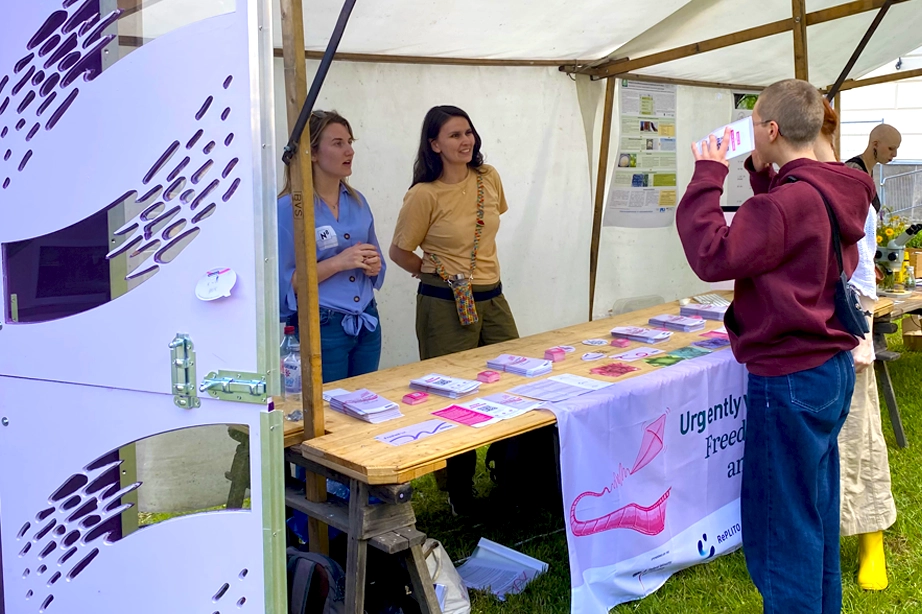 A booth at an outdoor event with two women standing behind a wooden table displaying various pamphlets and brochures. The booth has a white canopy and a banner on the front that reads "Urgently Wanted: Freedom of Research and Learning." A man wearing a maroon hoodie and blue jeans stands in front of the table, looking at the materials.