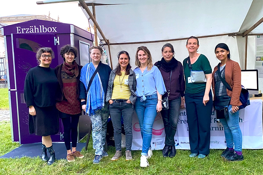 A group of seven women standing in front of a booth with a purple banner that reads "Erzählbox." The women are smiling and posing for the camera, with some standing on the grass and others in front of the booth. The booth has a white canopy and the banner includes additional text related to the event.