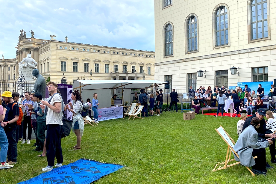 An outdoor gathering on a lawn with several people standing, sitting, and walking around. There are tents and booths set up around the perimeter, and a large historic building with arched windows in the background. Some people are interacting with the booths, while others are seated on the grass or in folding chairs, enjoying the event.