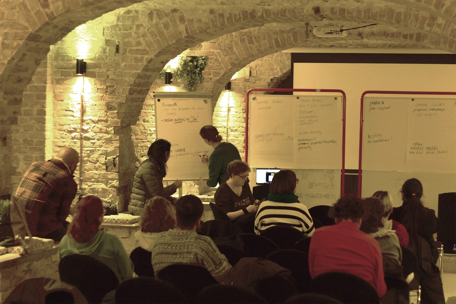 People seated in a brick-walled room, watching two individuals write on whiteboards covered with notes.