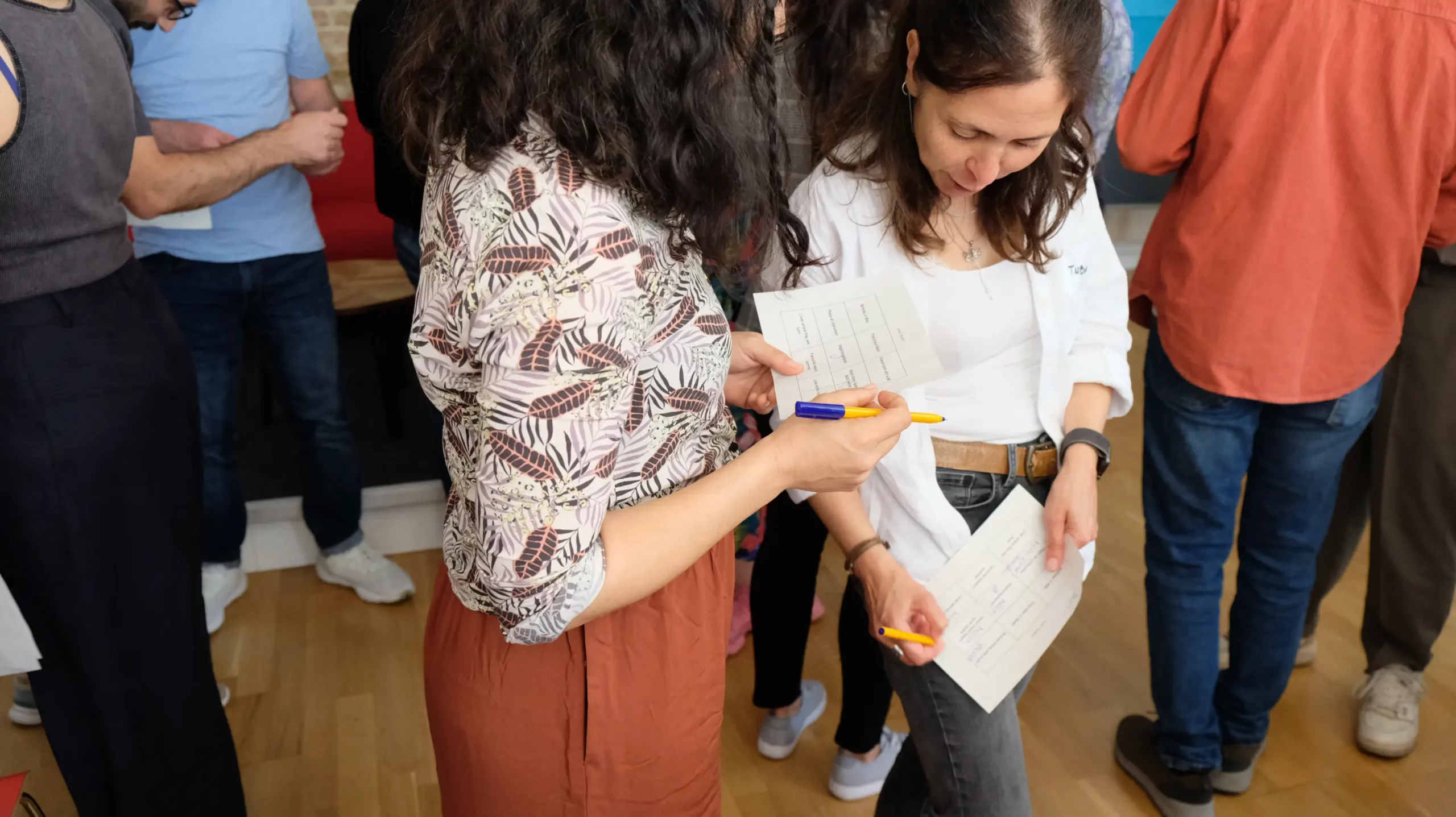 Two people standing close together, holding pens and papers, engaging in conversation while others in casual clothing interact in the background.