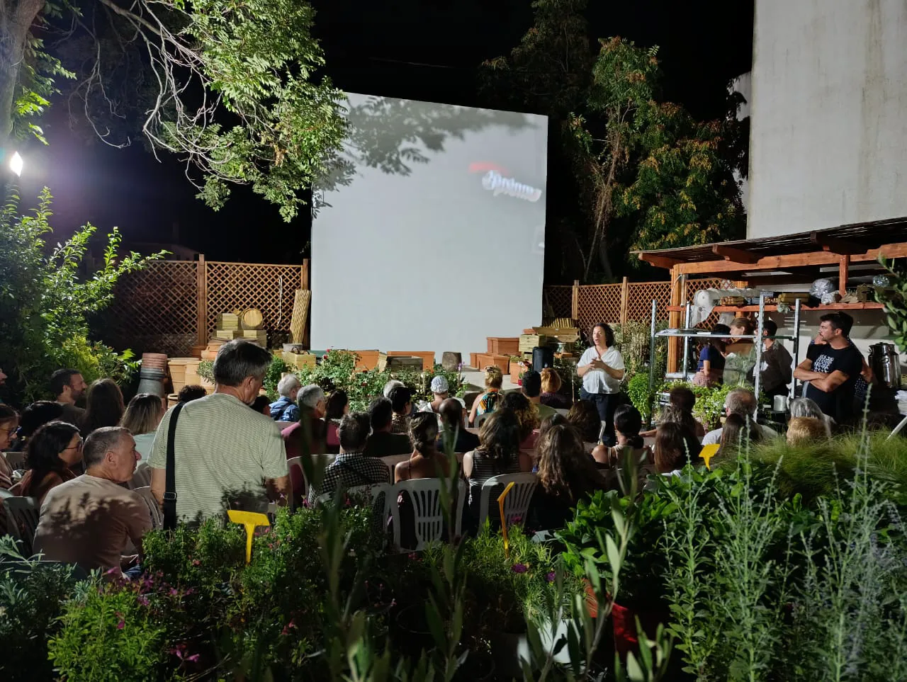 An outdoor nighttime gathering with people seated in front of a large projection screen, surrounded by greenery. A woman is speaking to the audience.